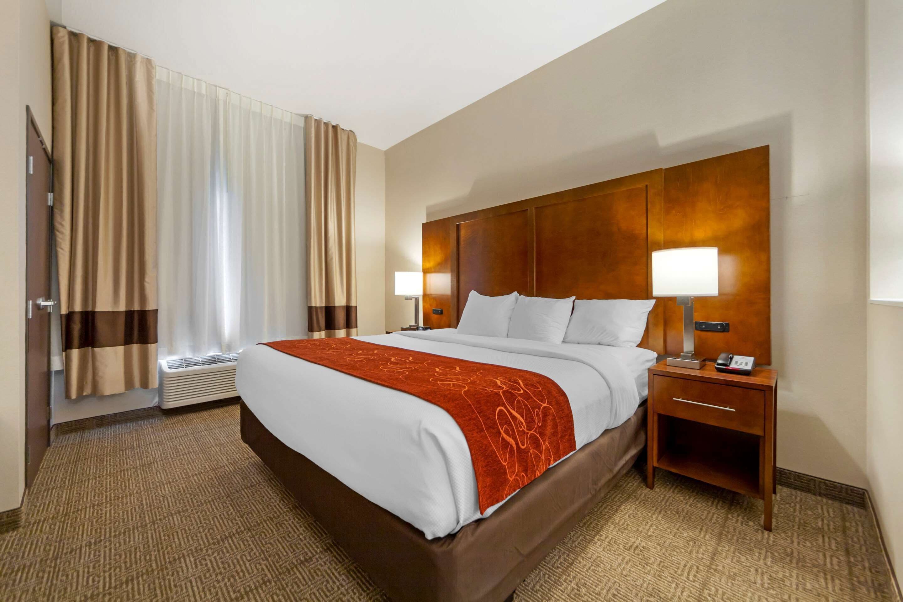 A hotel room with a large bed, two bedside tables with lamps, a window with curtains, and a wall-mounted heater.