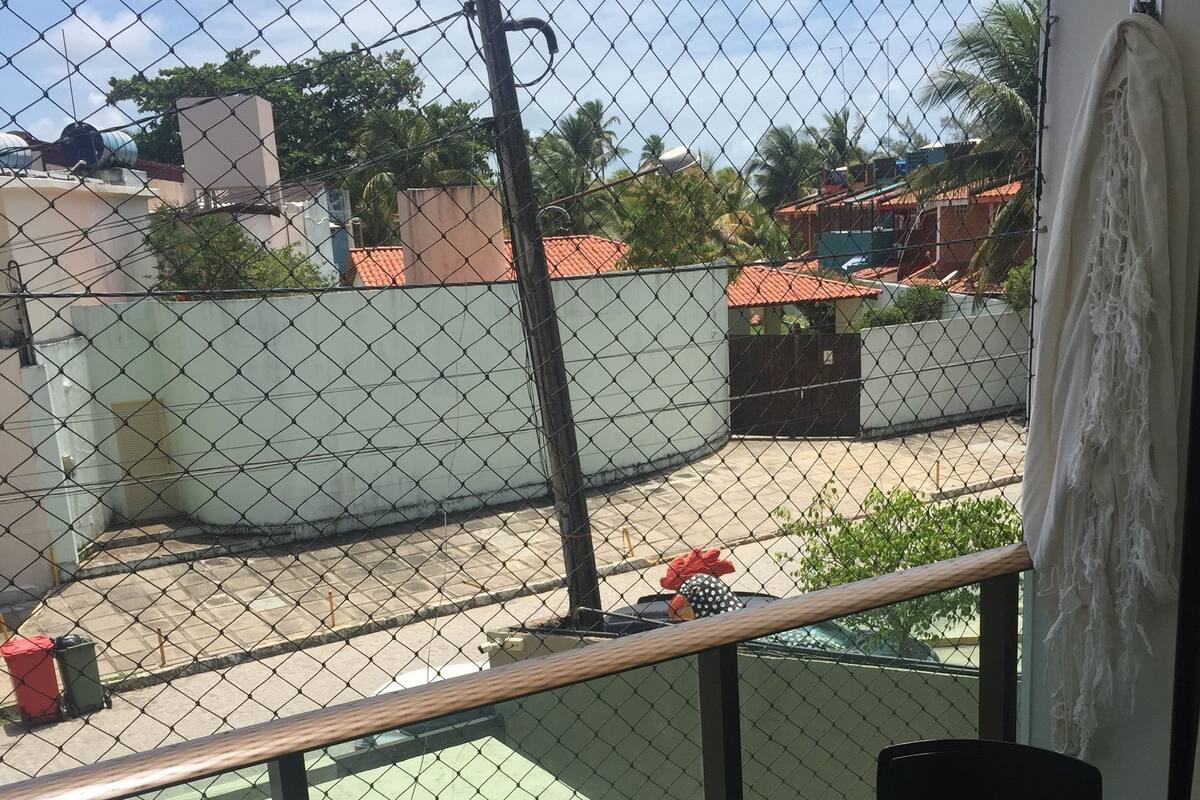 A balcony with a table and two chairs, a chain-link fence, and a view of the surrounding neighborhood.