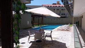 Une piscine avec des chaises longues et un parasol, entourée d’une terrasse en briques et d’un bâtiment en arrière-plan.