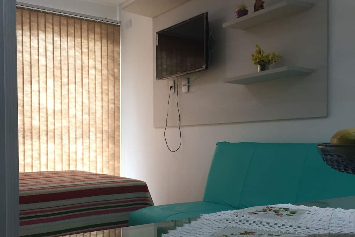 A room with a bed, a TV mounted on the wall, a sofa, and a table with a tablecloth.