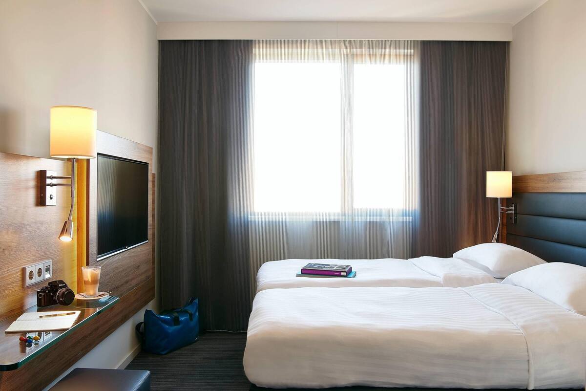 Ein Hotelzimmer mit einem Bett, einem Schreibtisch mit Lampe, einem Fernseher und einem Fenster mit Vorhängen.