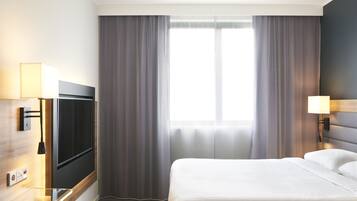 Habitación de hotel con una cama grande, un televisor de pantalla plana, una mesita de noche con lámpara y un ventanal con cortinas.