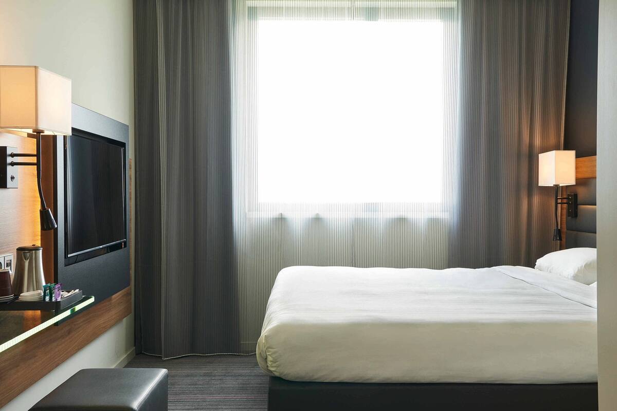 Ein Hotelzimmer mit Bett, Fernseher, Schreibtisch mit Lampe und einem Fenster mit Vorhängen.
