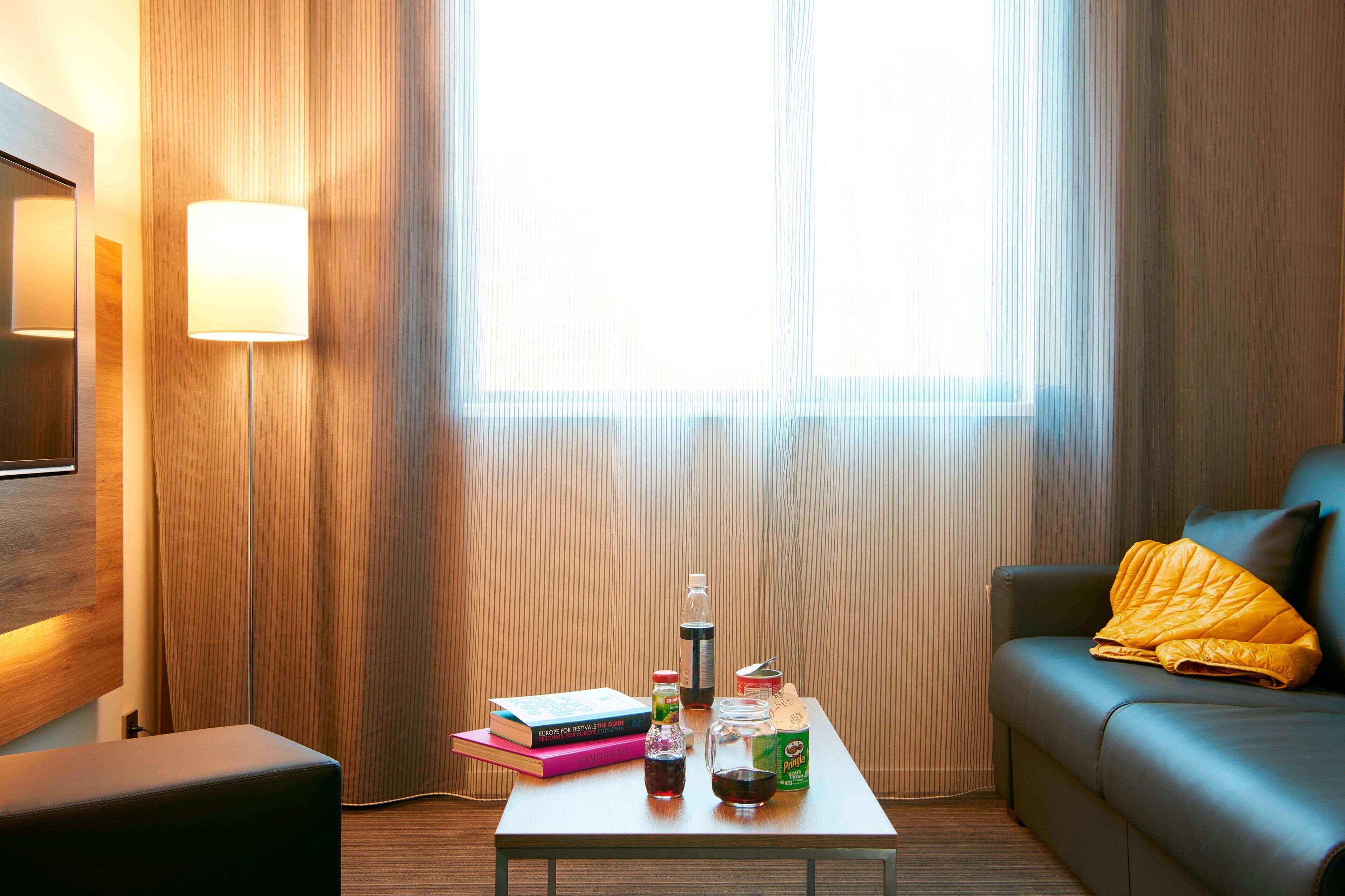 Una habitación de hotel moderna con piso de madera, un sofá de cuero negro, una mesa de centro de vidrio y un ventanal con cortinas de gasa.