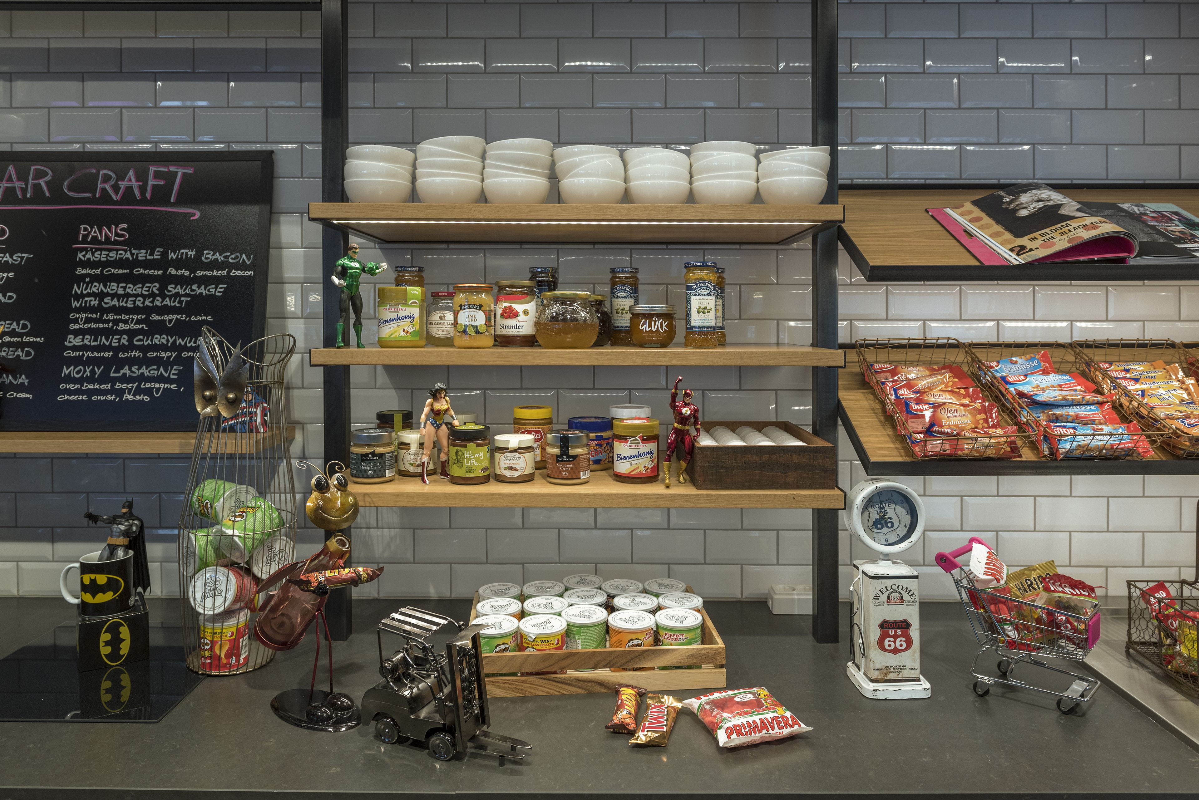 A kitchen with shelves stocked with jars of condiments, a display of Batman-themed items, and a shopping cart with snacks.