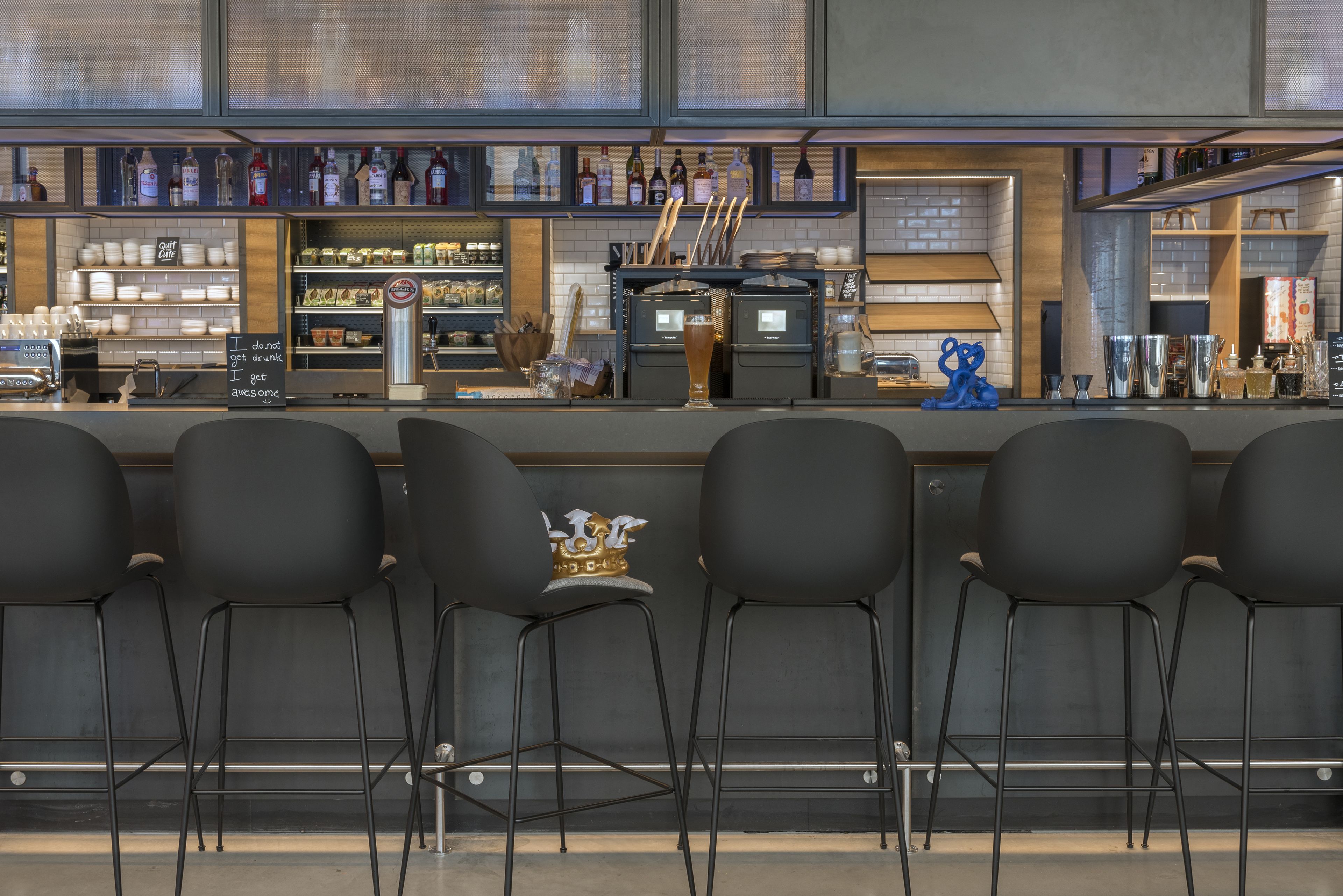 Una zona de bar con mostrador, taburetes y estanterías llenas de botellas y vasos.