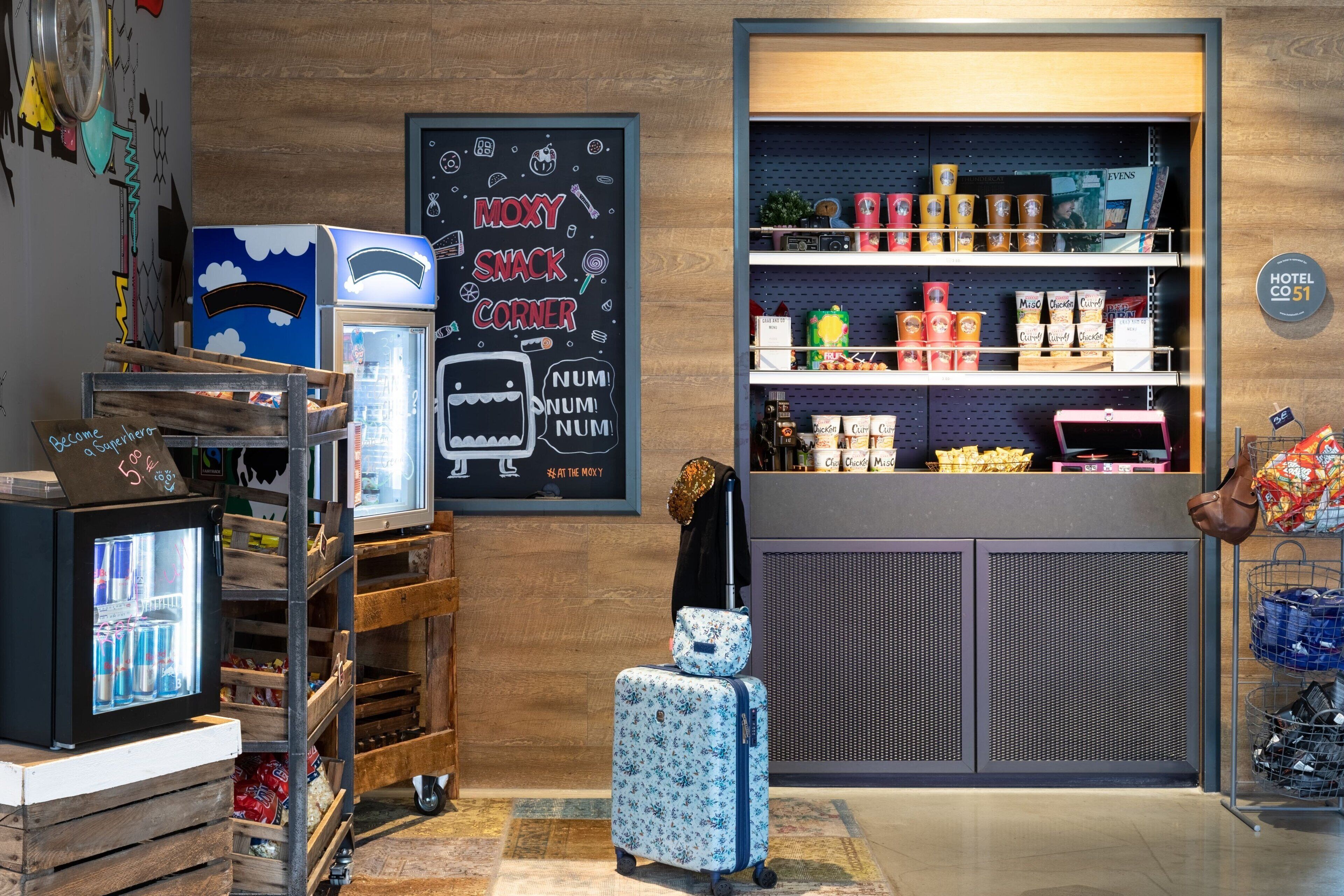 Un coin accueillant avec un panneau noir où est écrit « MOXY SNACK CORNER », un distributeur automatique et une sélection de collations et de boissons présentées sur des étagères.