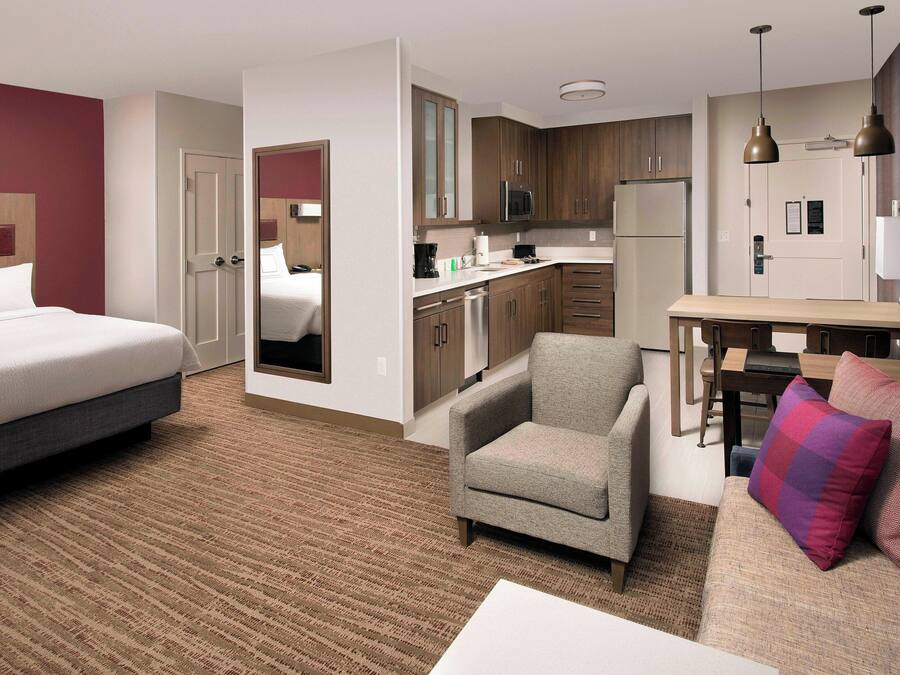 Habitación de hotel con cama, sofá, sillón, mesita, zona de cocina y espejo.