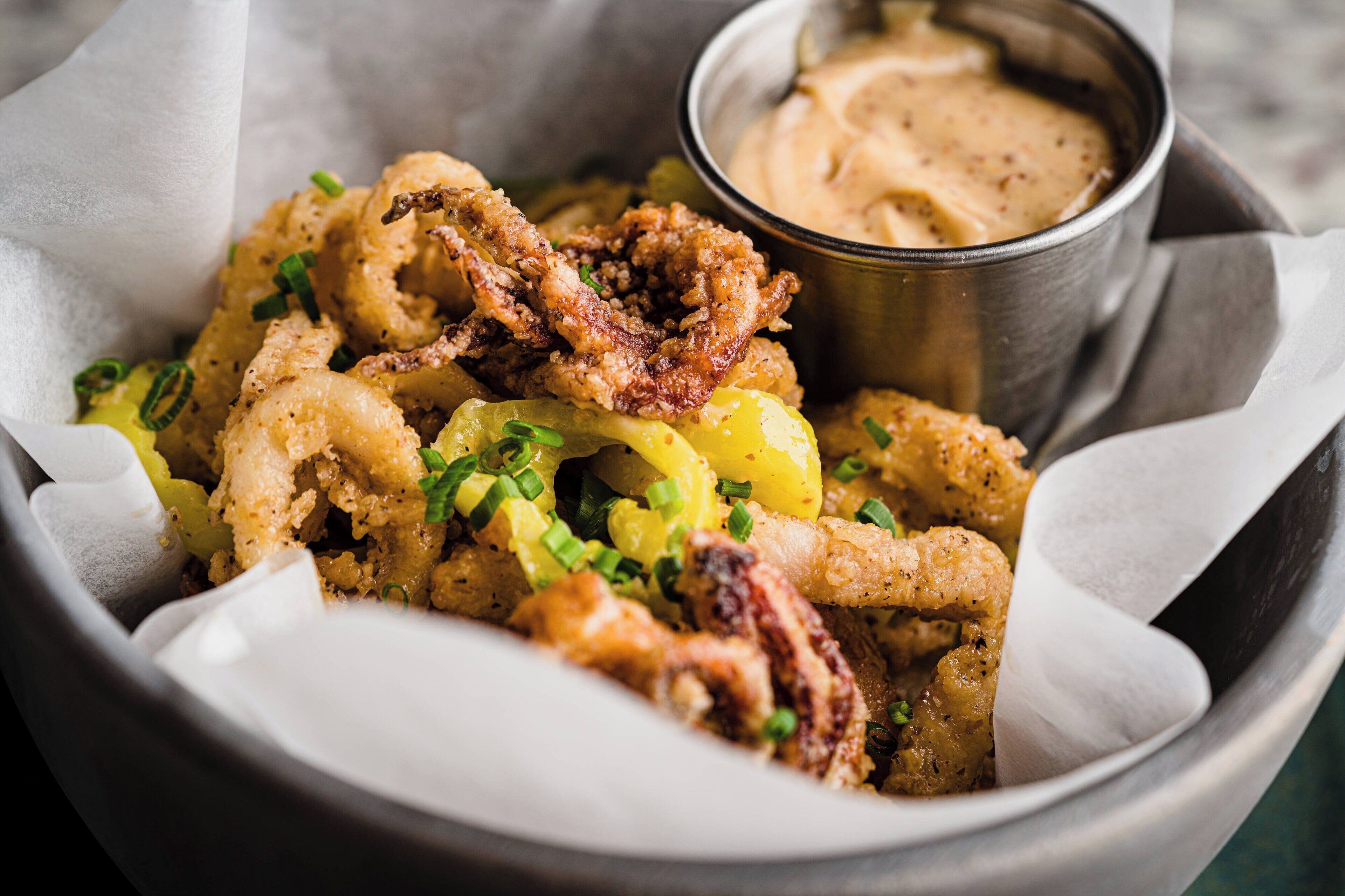 Un bol de calamares fritos con gajos de limón y cebolletas, acompañado de una pequeña taza de salsa para mojar.
