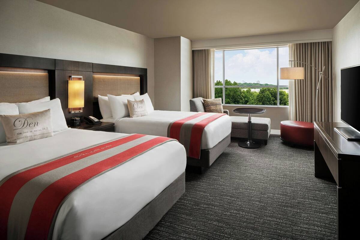 A hotel room with two beds, a desk, a chair, a TV, and a view of the outdoors.