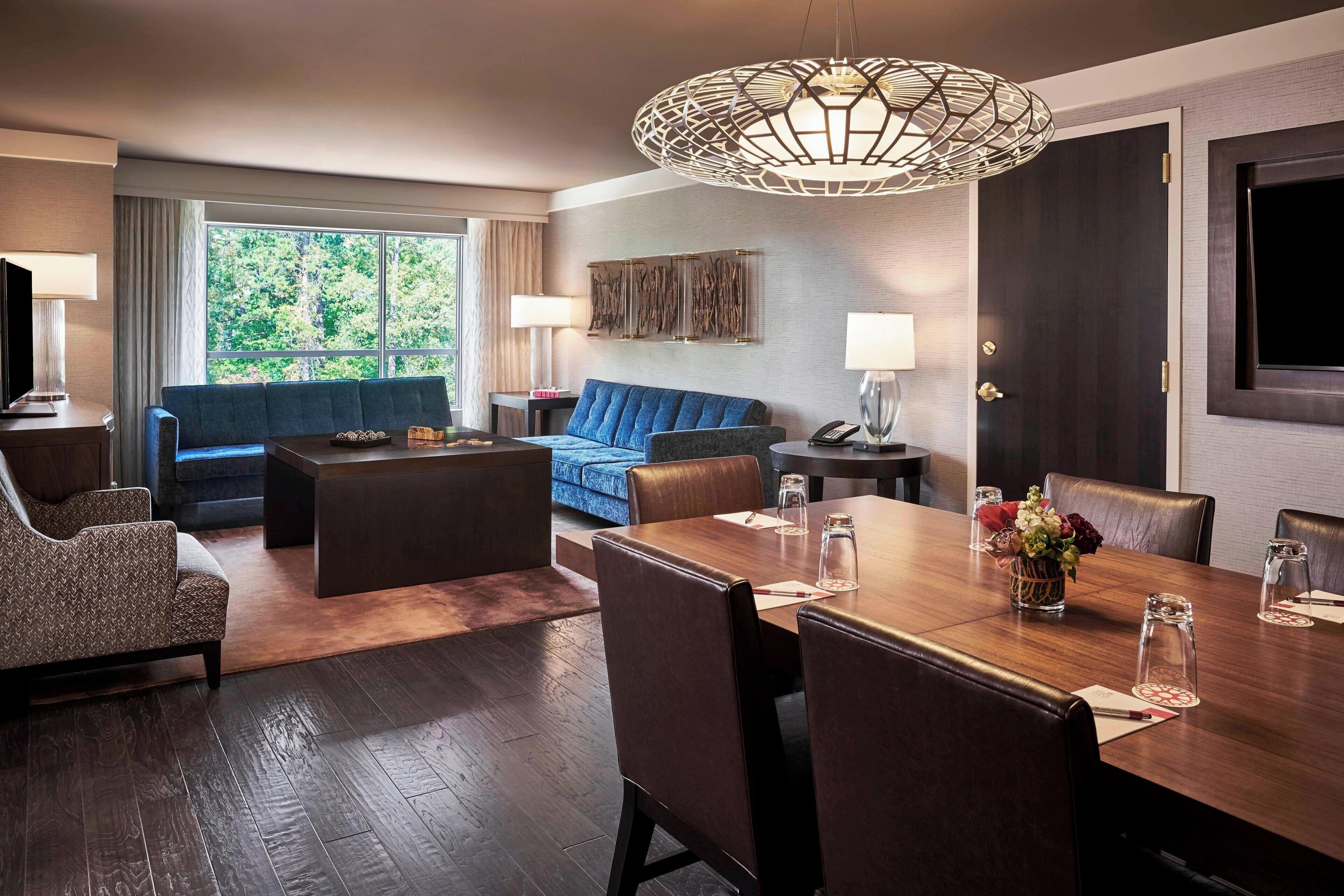 Habitación de hotel con zona de comedor, sofá, televisor y un amplio ventanal con vistas a árboles.