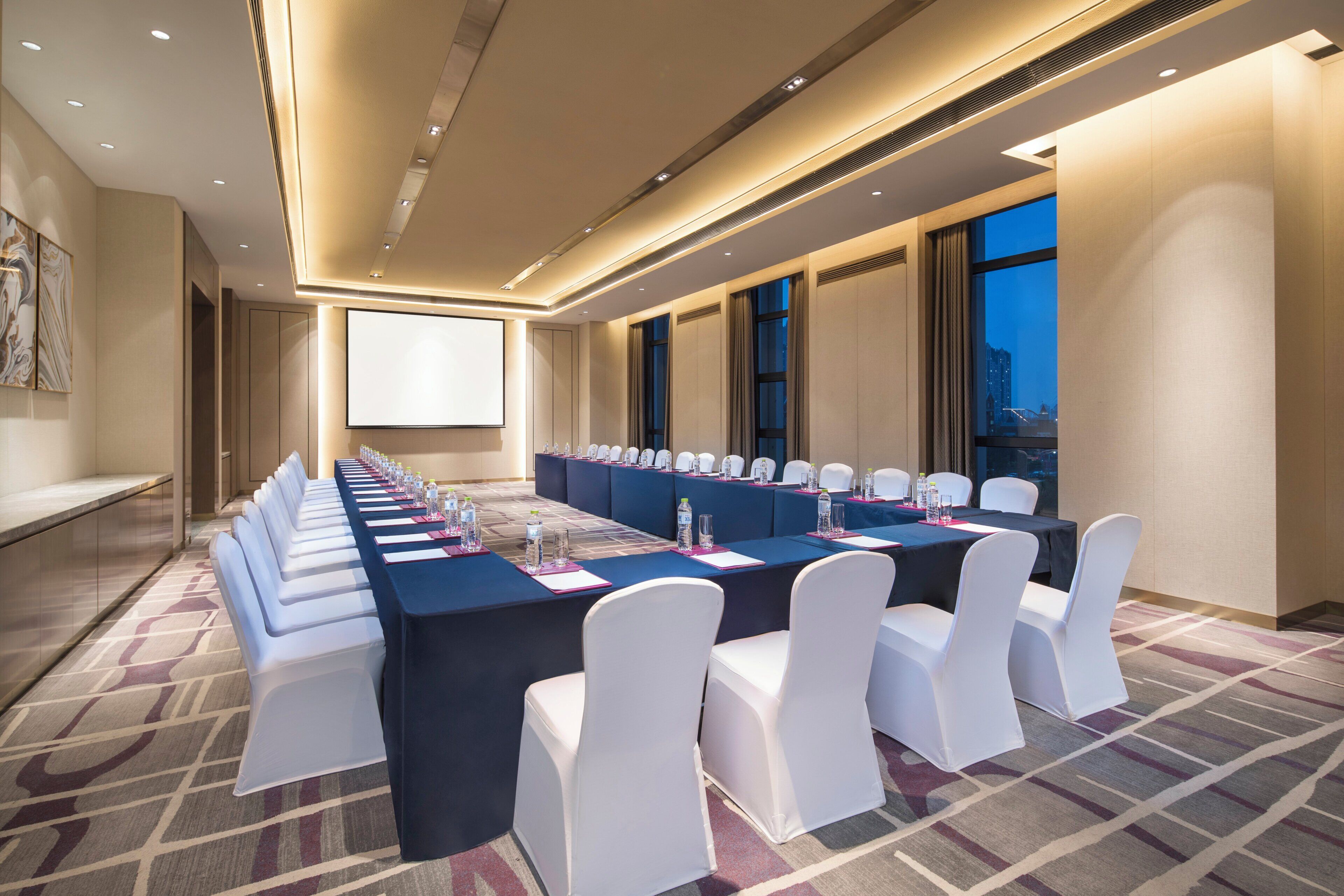 A conference room with a long table, white chairs, and a projector screen.