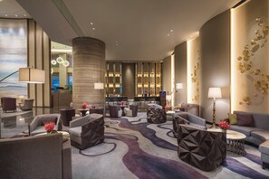 Un vestíbulo de hotel con una mesa de recepción, una zona de estar con sofás y sillones, un pilar grande y una pared decorativa con diseños circulares.