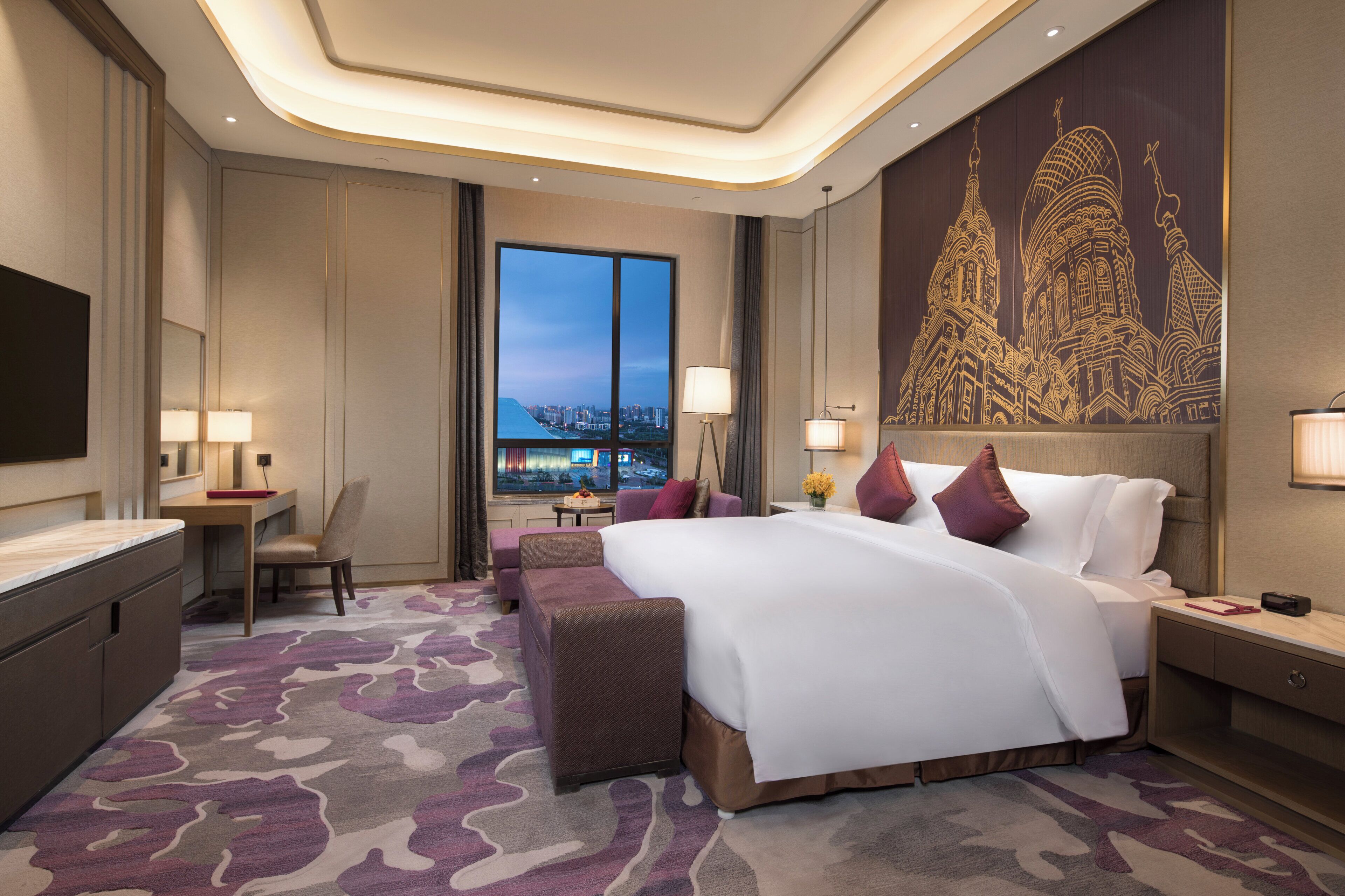 A hotel room with a large bed, a desk, a chair, and a view of a cityscape through the window.