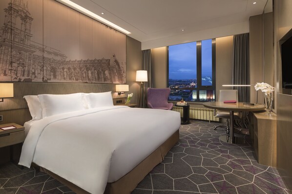 Habitación de hotel con una cama grande, un escritorio, una silla, vistas de la ciudad al anochecer y un mural grande en la pared.