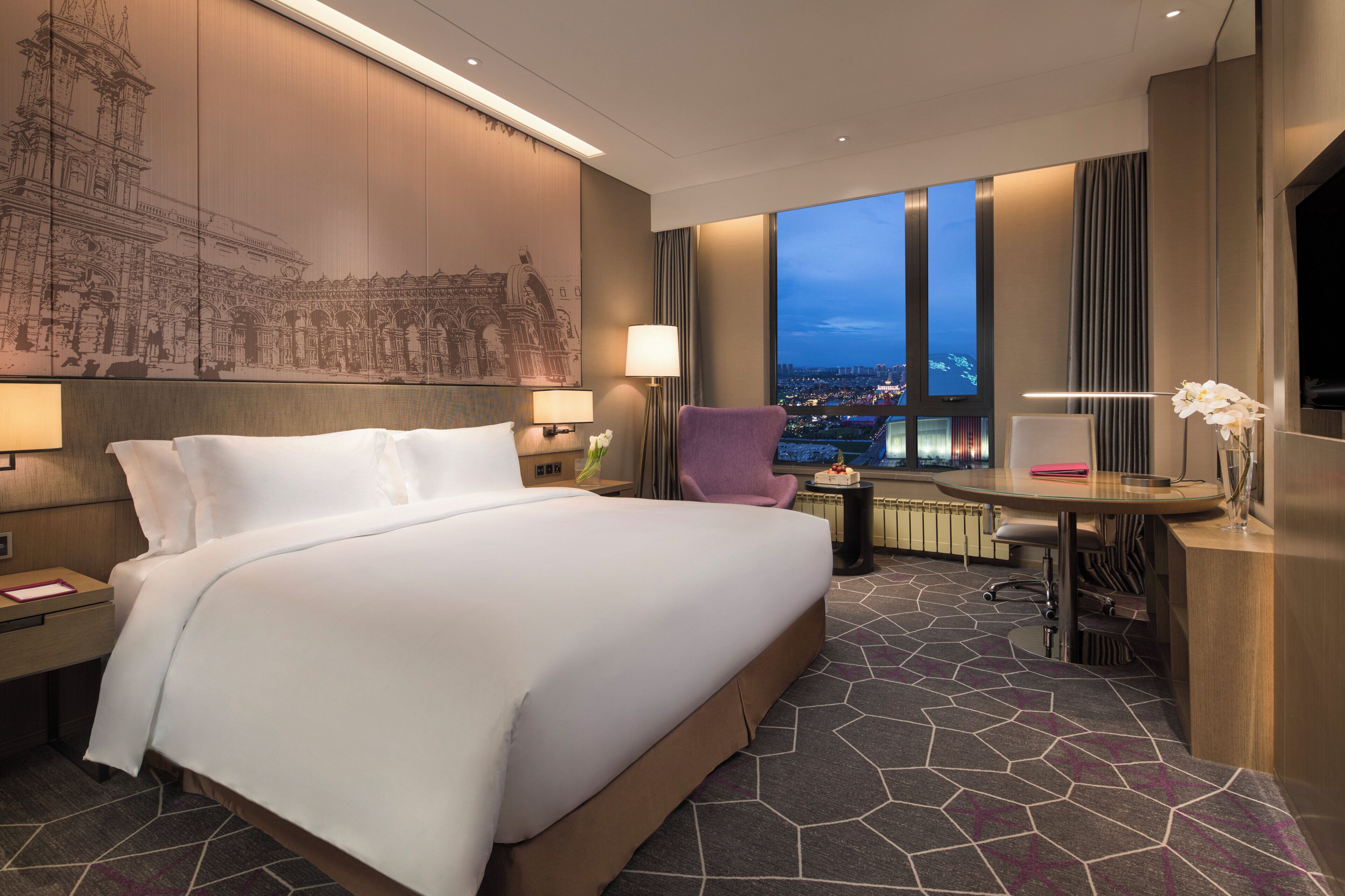 A hotel room with a large bed, a desk, a chair, a view of the cityscape, and a decorative wall.