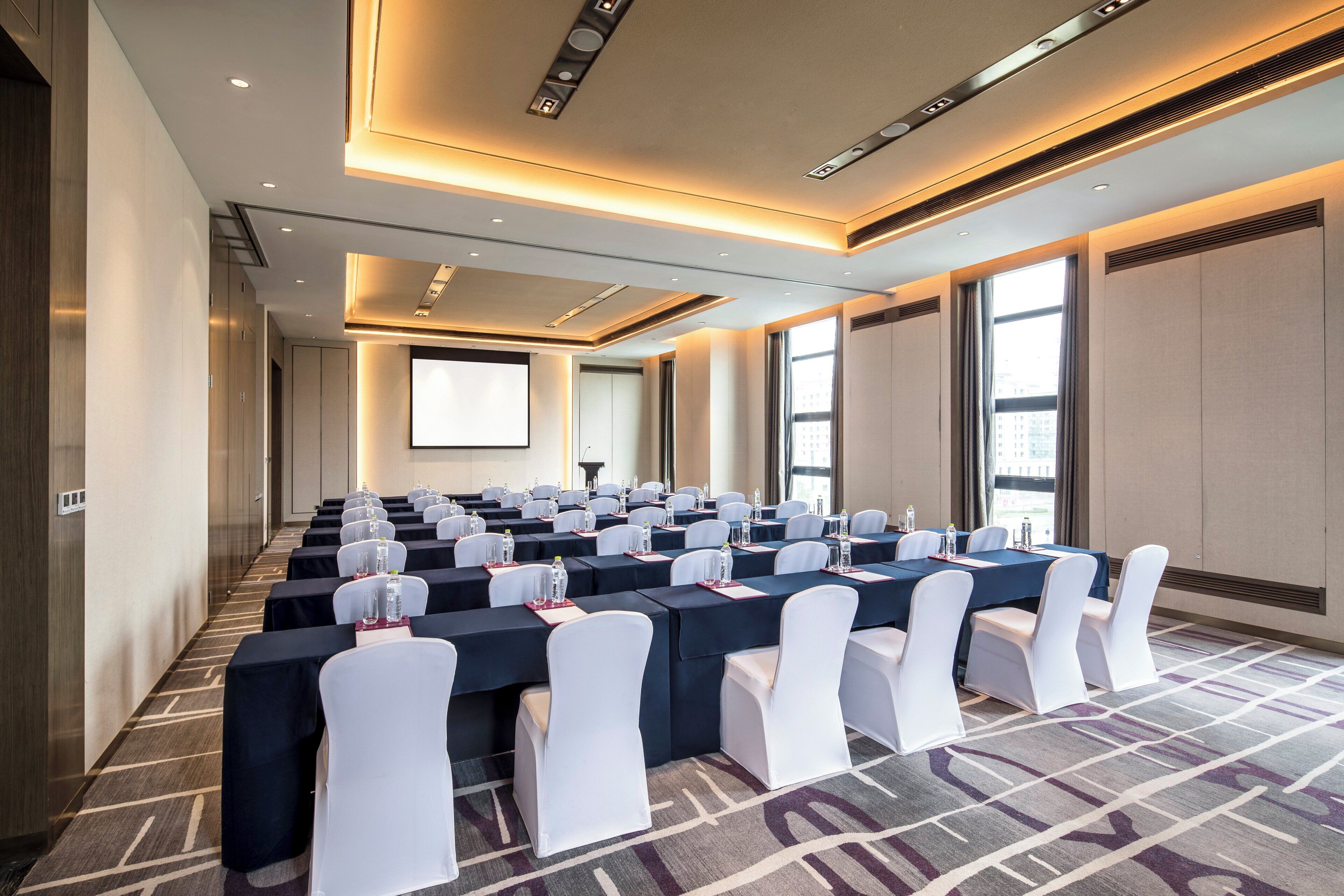 Un salón de conferencias con filas de mesas, sillas blancas y una pantalla grande.