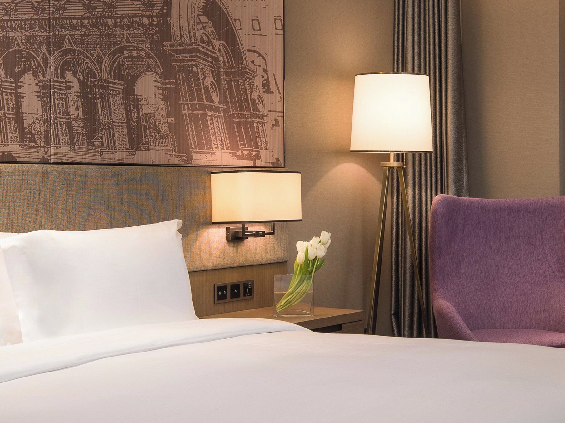 Una habitación de hotel con una cama, una mesita de noche con una lámpara, una silla y un mural decorativo en la pared.