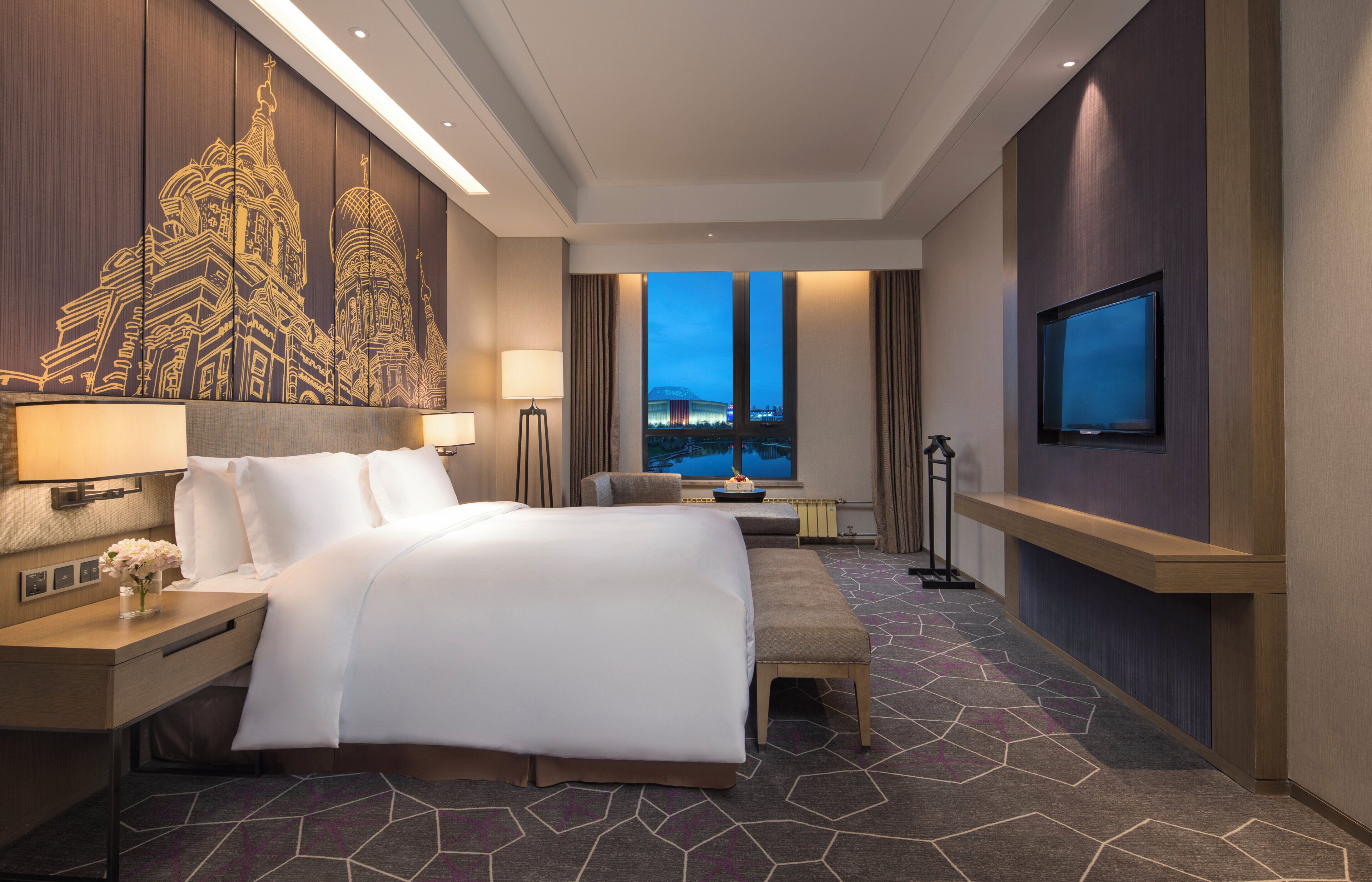 Habitación de hotel con una cama grande, un escritorio, un televisor y vistas a un paisaje urbano al atardecer.