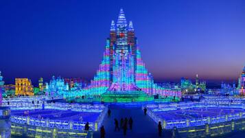 A large, illuminated ice sculpture with a central spire, surrounded by smaller ice structures and a cityscape in the background.