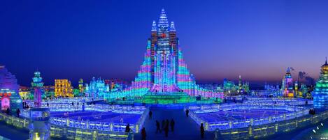 A large, illuminated ice sculpture with a central spire, surrounded by smaller ice structures and a cityscape in the background.