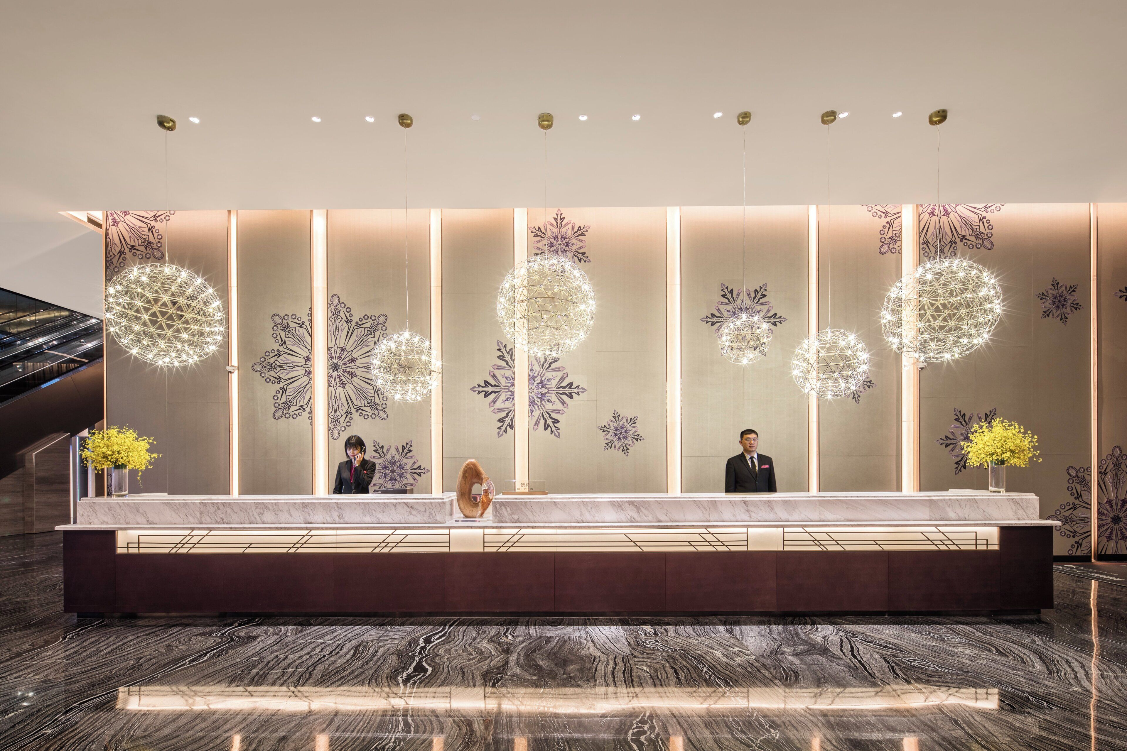 A hotel reception desk with a marble counter, decorative wall panels, and a modern chandelier.