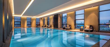 A modern indoor swimming pool with clear blue water, surrounded by lounge chairs and large windows offering city views.