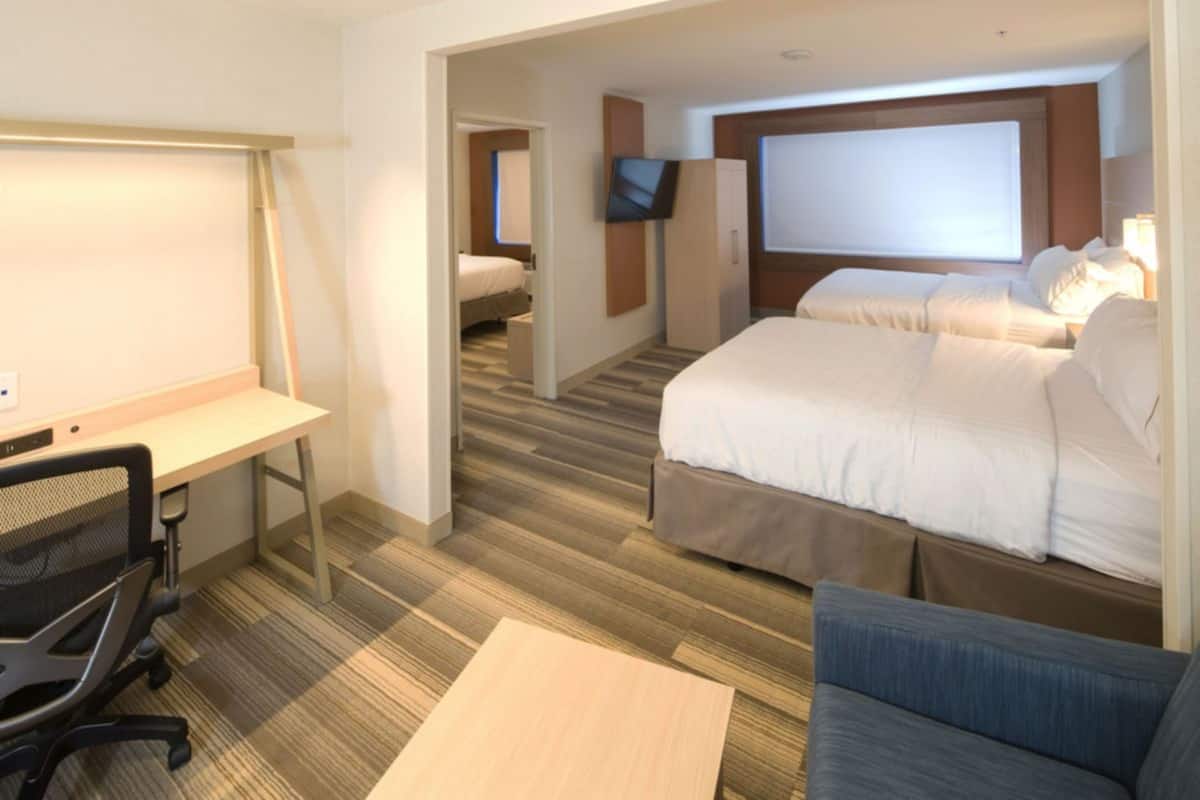 Una habitación de hotel con escritorio, dos camas, televisor y un ventanal grande.