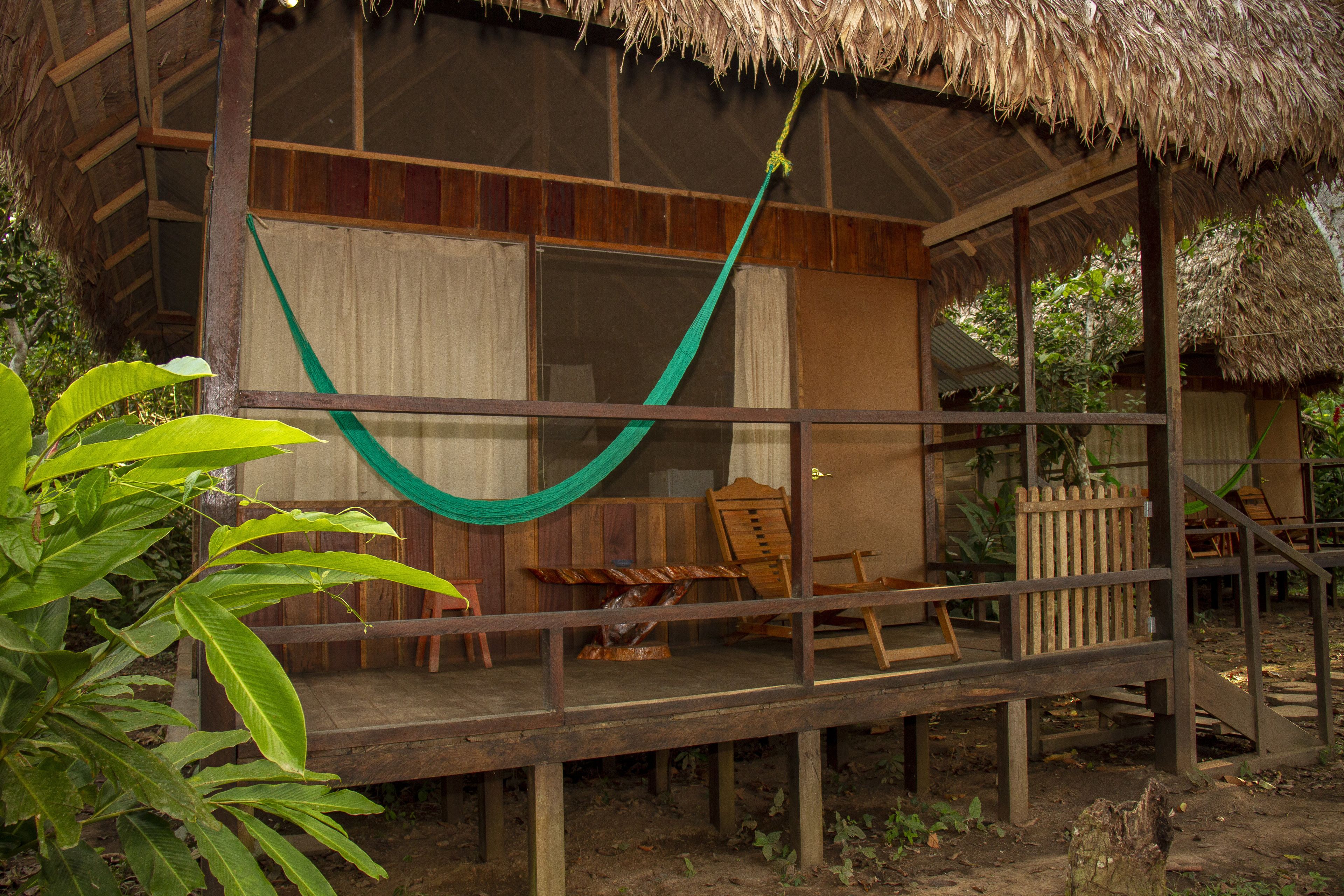 A wooden structure with a thatched roof, a hammock, and a wooden deck with chairs and a table.