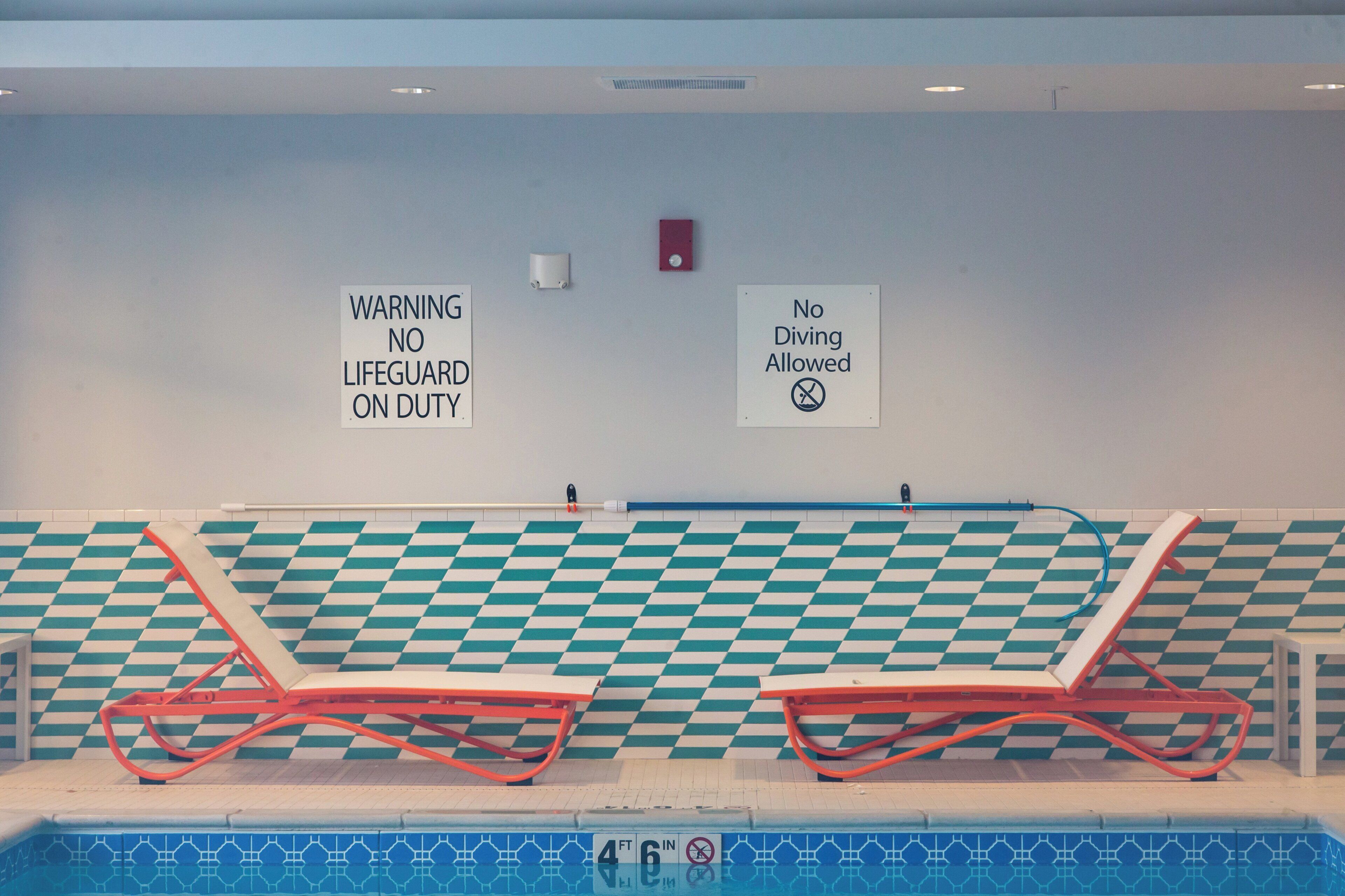 A swimming pool with two red lounge chairs and two warning signs on the wall.