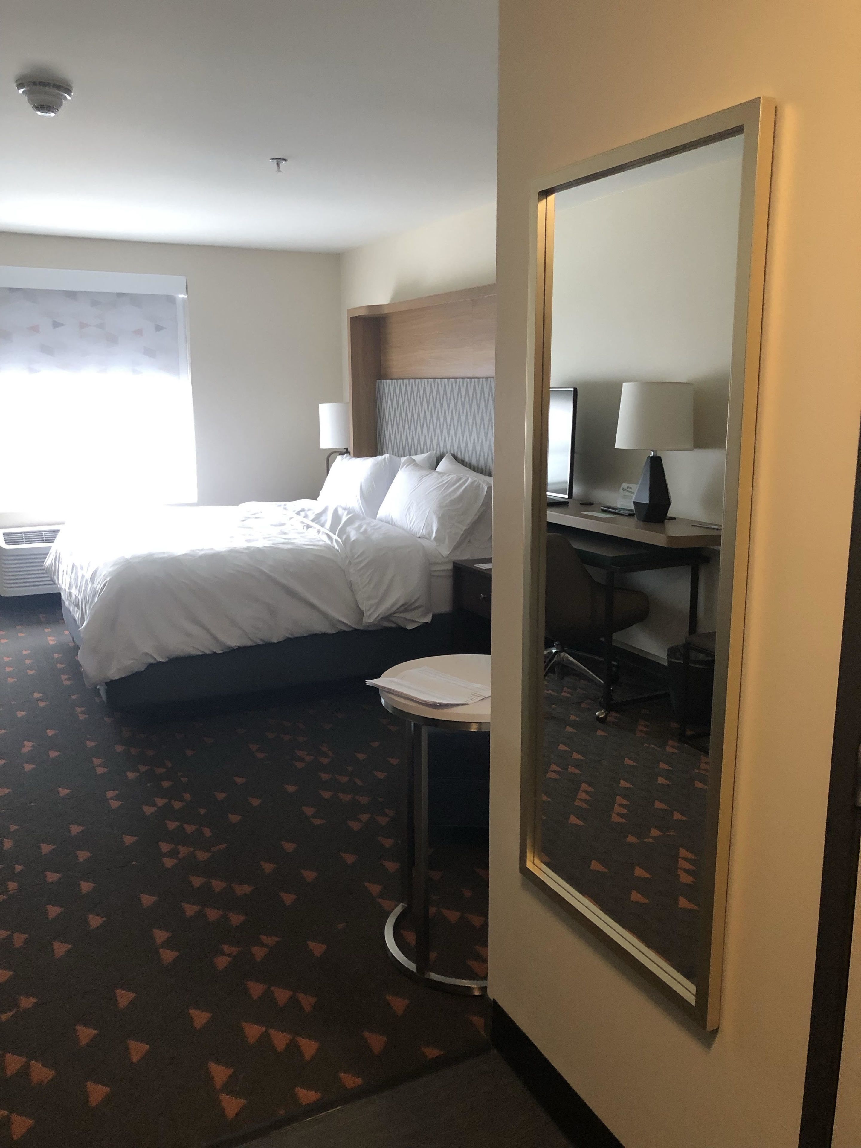A hotel room with a bed, bedside table, lamp, and a large mirror.