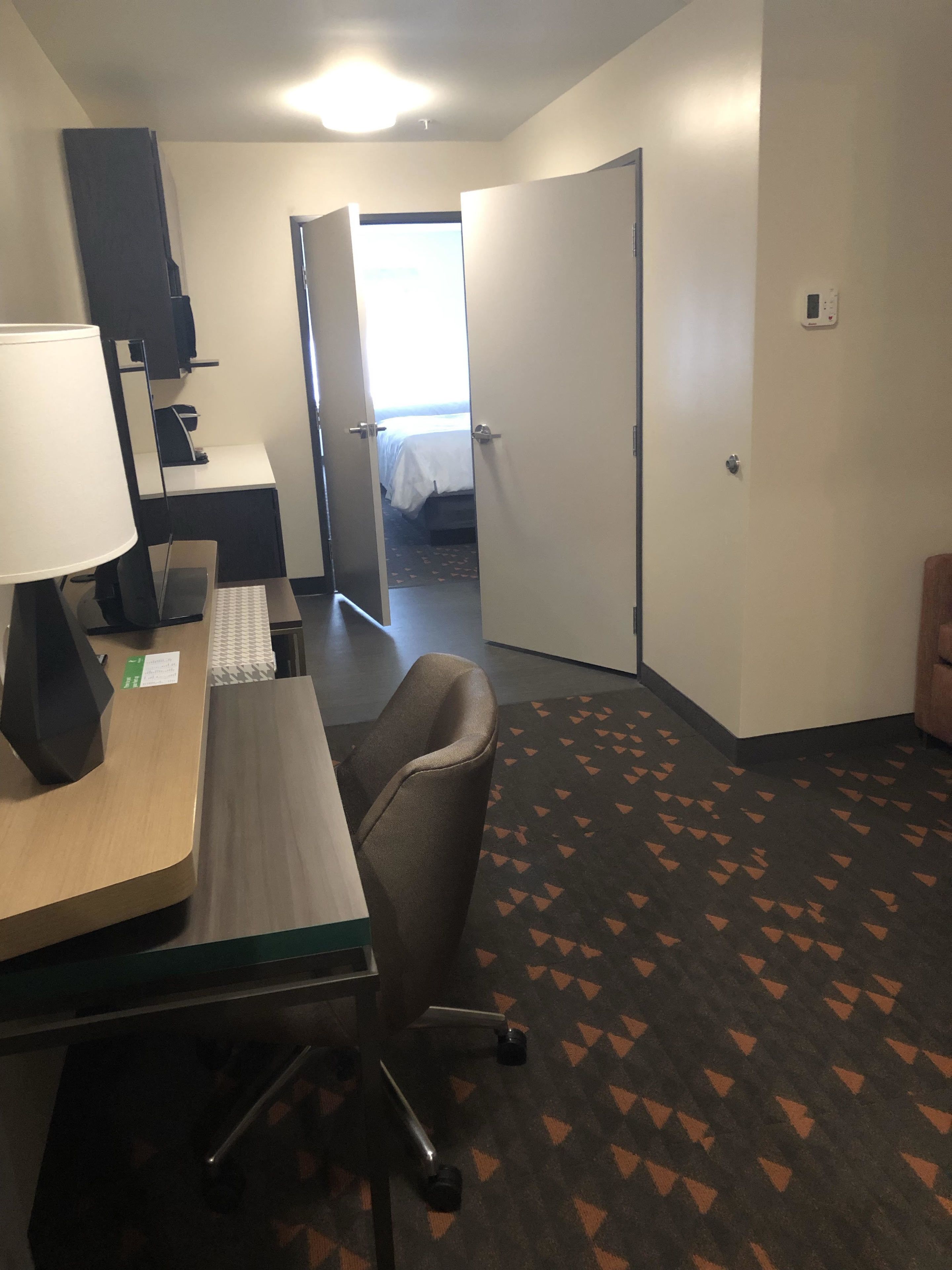 A hotel room with a desk, chair, lamp, and a bed in the background.