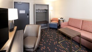 Habitación de hotel con sofá, una silla, una mesita, televisión, lámpara, puerta y zona de kitchenette.