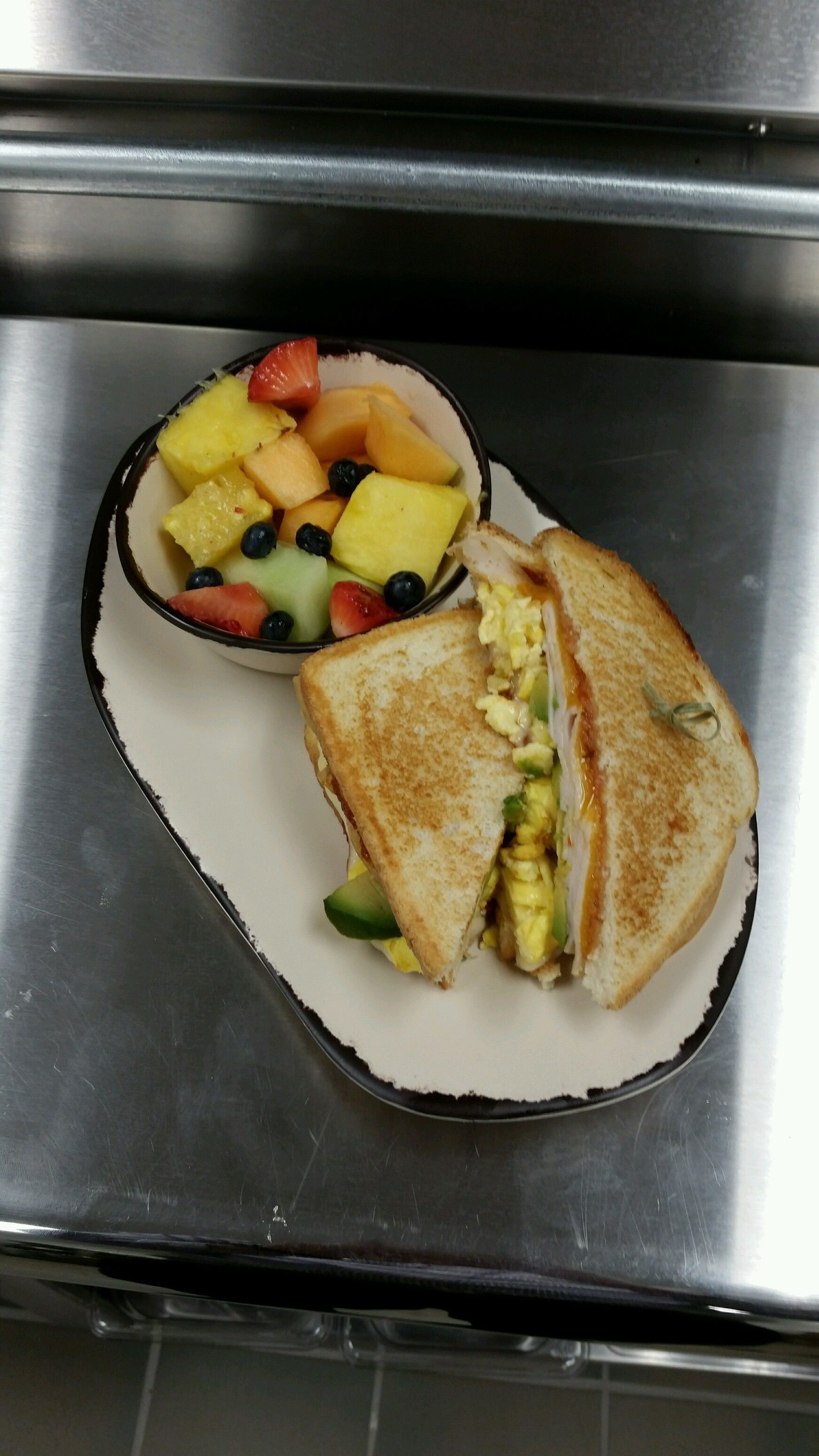 A sandwich cut in half with avocado and egg, and a bowl of fruit including strawberries, blueberries, and pineapple.