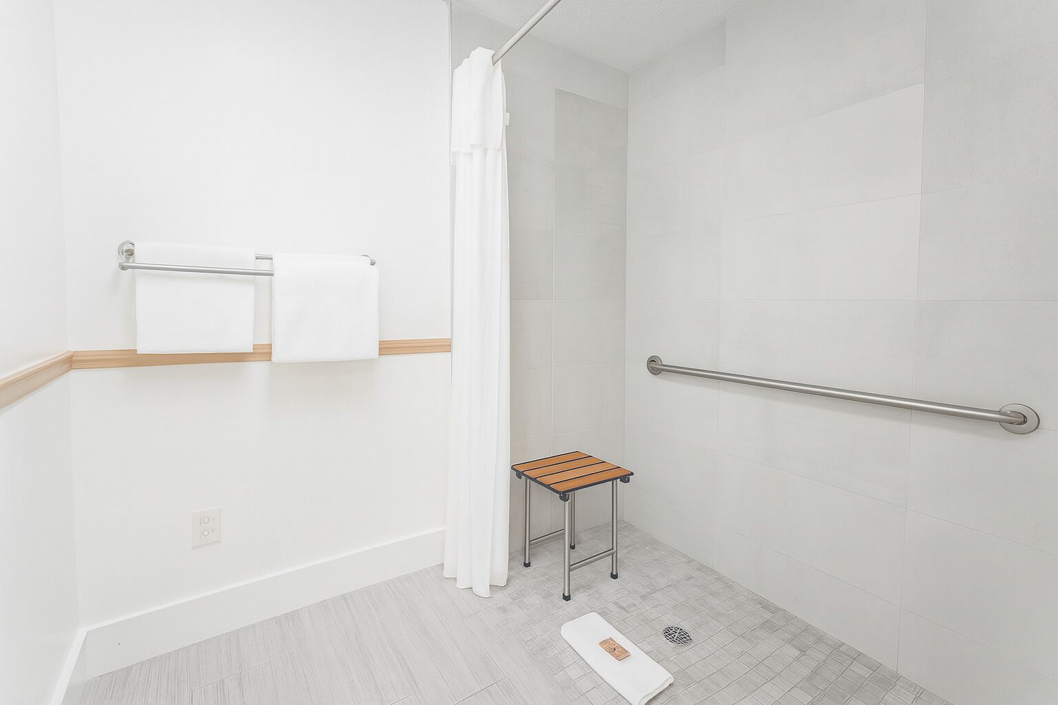 A shower area with a white curtain, a grab bar, a small wooden stool, and a towel rack with two towels.