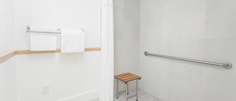 A shower area with a white curtain, a grab bar, a small wooden stool, and a towel rack with two towels.