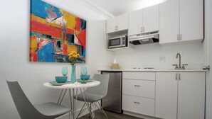 A modern kitchen with white cabinets, a stainless steel dishwasher, and a microwave. There is a dining area with a white table and two gray chairs. A colorful abstract painting is on the wall above the table. - Spinnaker Inn of Naples (Naples)