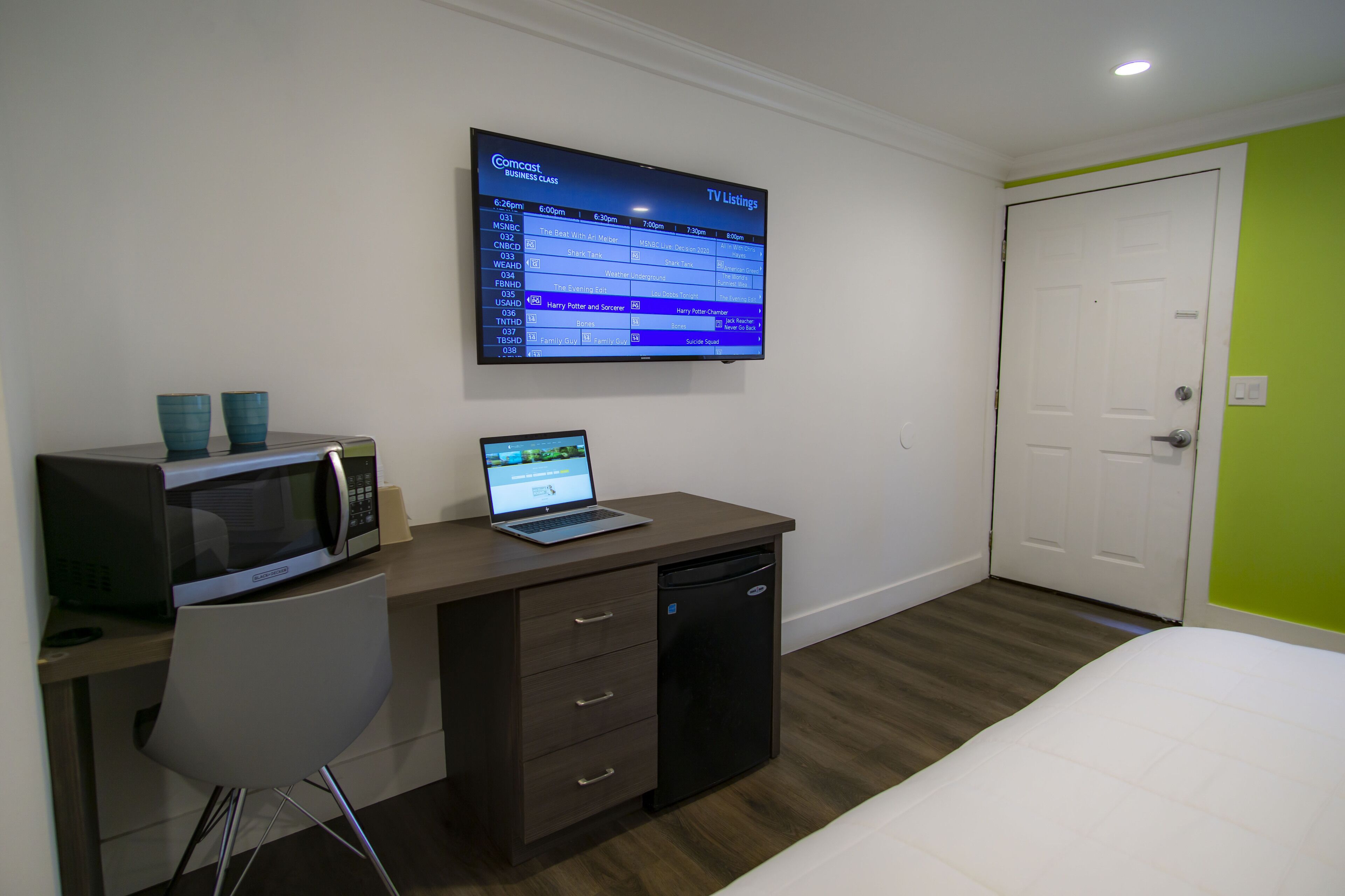 A hotel room with a bed, a desk with a laptop, a microwave, and a TV displaying TV listings.