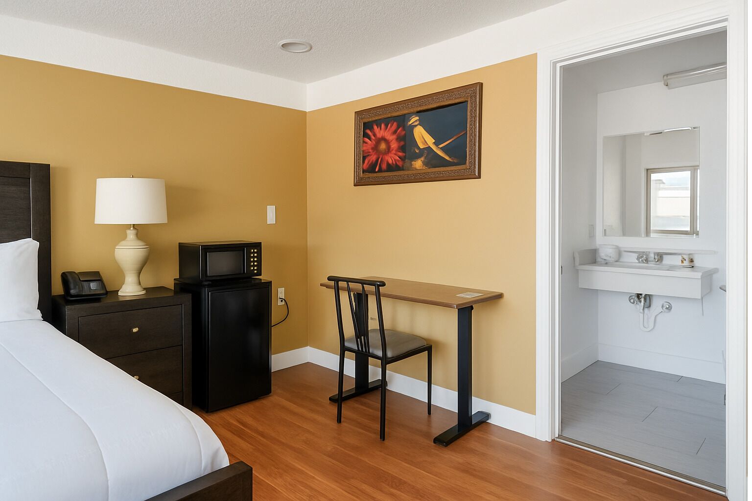 A hotel room with a bed, a desk with a lamp, a microwave, a chair, a painting, and a bathroom with a sink.