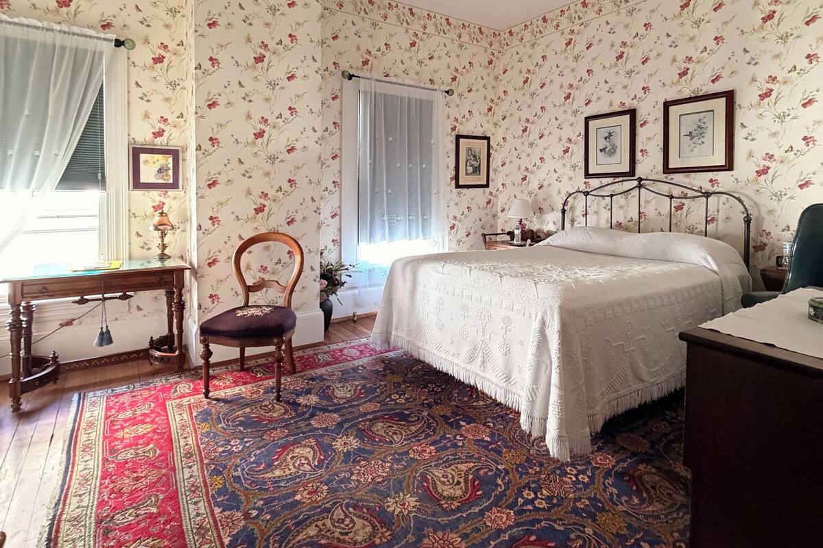 Een slaapkamer met bloemenbehang, een bed met een witte dekbedovertrek, een houten stoel, een bureau met een lamp en een plafondventilator.