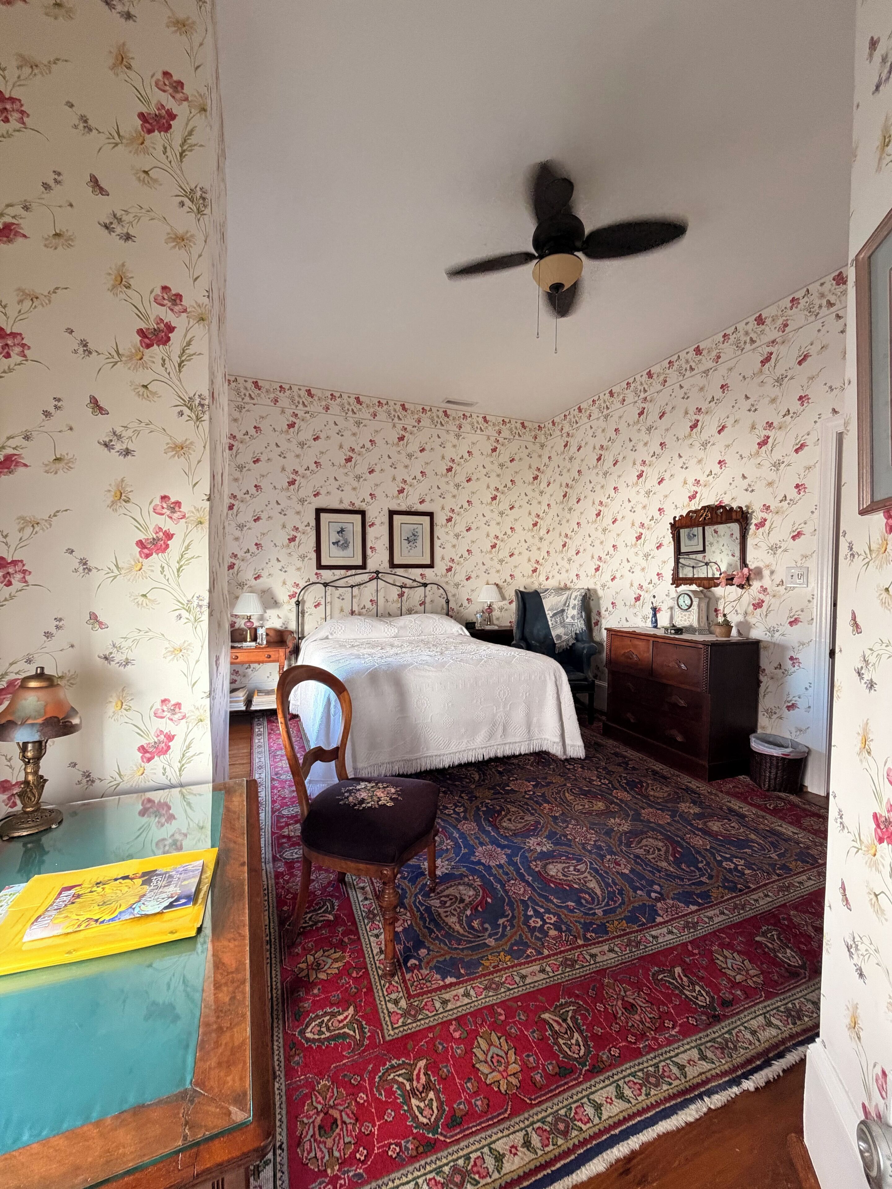Une chambre à coucher avec un papier peint à motifs floraux, un lit, une chaise, un bureau, un ventilateur de plafond et une commode.