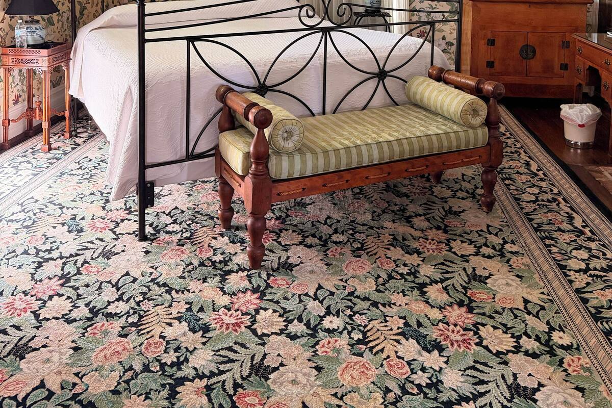 Een slaapkamer met een hemelbed, een houten bankje, een kroonluchter, een spiegel en bloemenbehang.