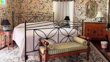 Ein Schlafzimmer mit einem Himmelbett, einer Holzbank, einer Chandelüster, einem Spiegel und blumigem Wandpapier.