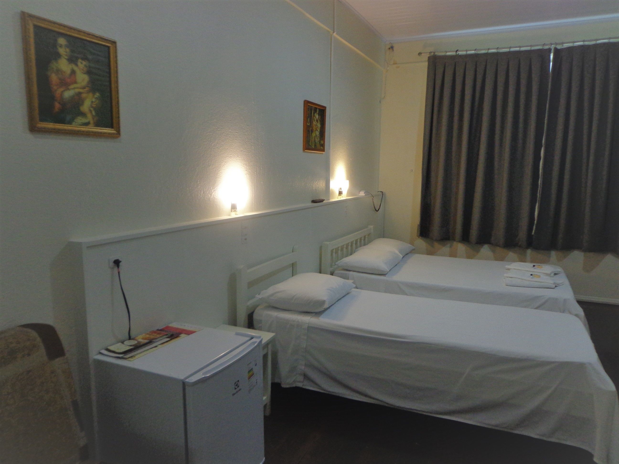 Quarto de hotel com duas camas de solteiro, um pequeno frigorífico, uma escrivaninha e uma janela com cortinas.