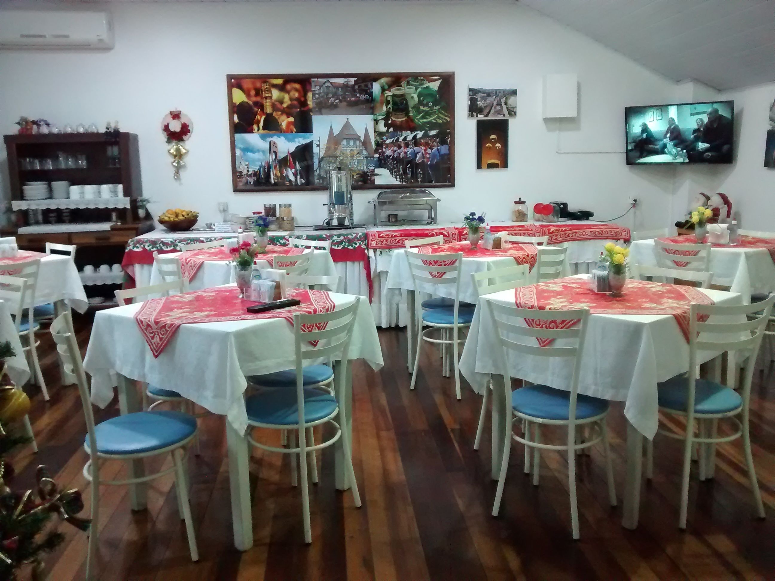Uma sala de jantar com várias mesas cobertas por toalhas brancas e centros de mesa vermelhos, cadeiras de madeira e uma televisão presa à parede.