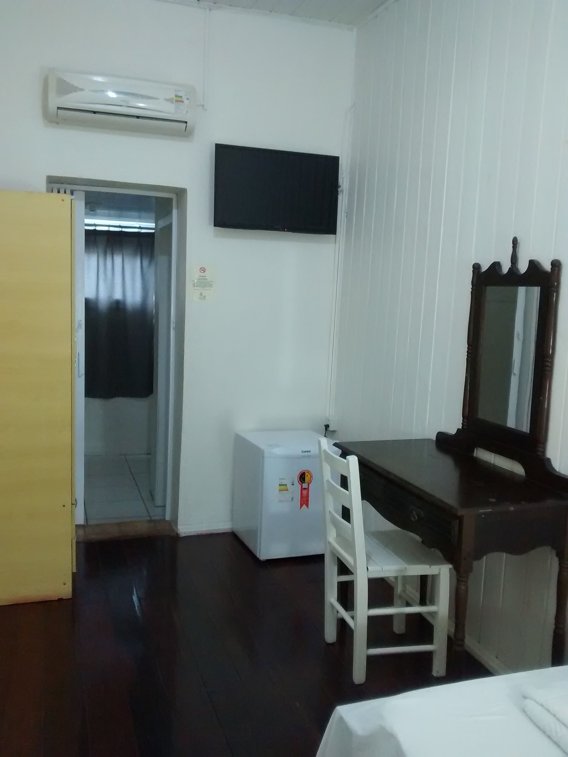 Quarto de hotel com cama, escrivaninha com cadeira, televisão fixada na parede e um pequeno frigorífico.