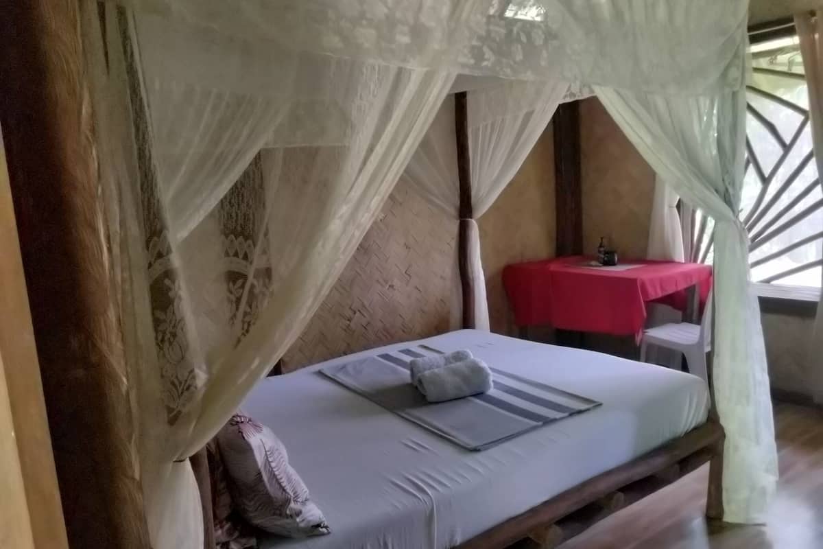 Un lit simple avec un baldaquin, une petite table recouverte d’une nappe rouge et une chaise.