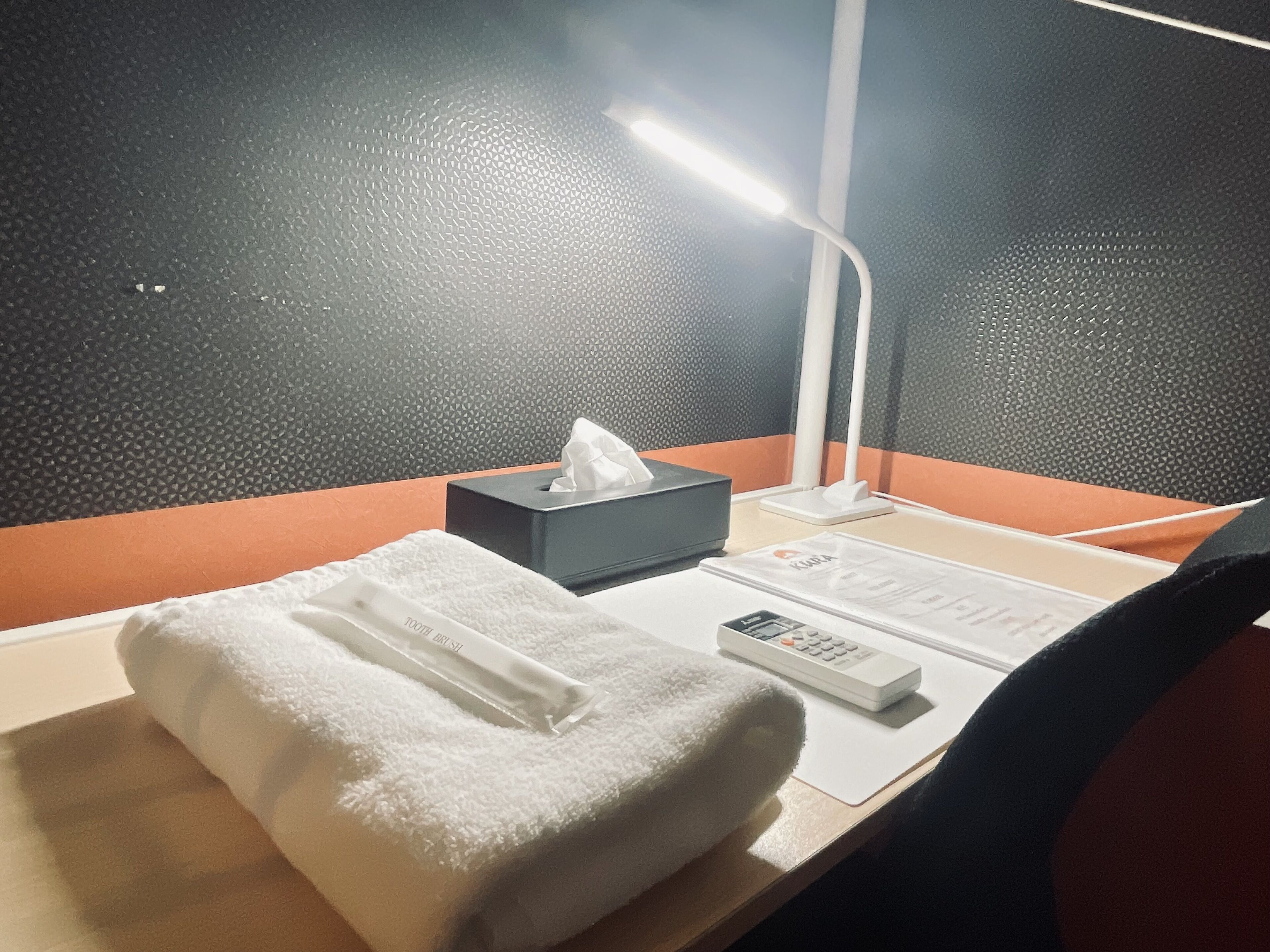A hotel room with a bed, a bedside table, a lamp, a tissue box, a remote control, and a wall with a textured pattern.
