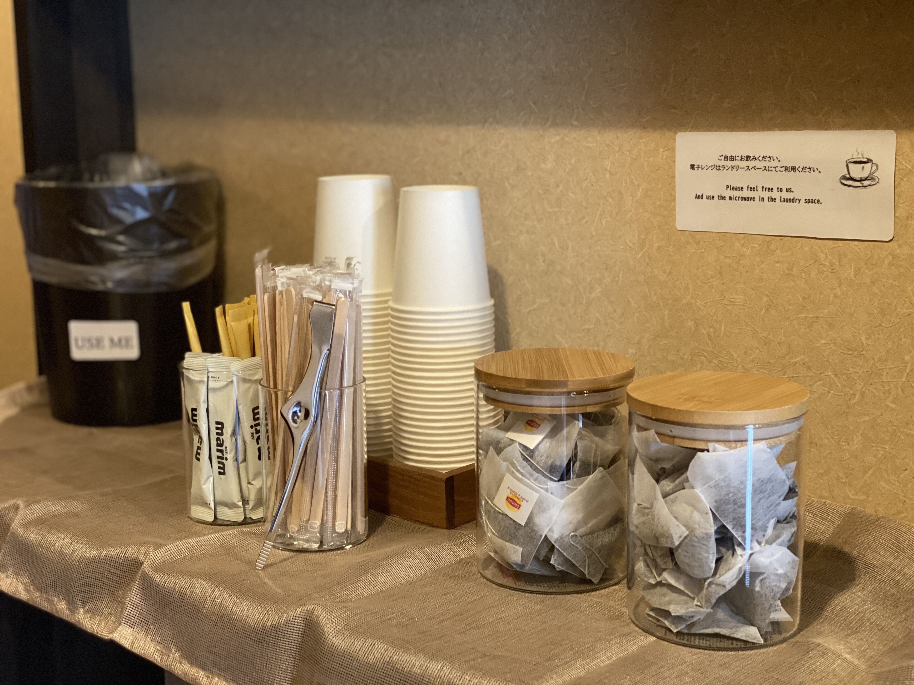 A table with stacks of white cups, a trash bin labeled "USE ME", and two glass jars with lids containing tissues.