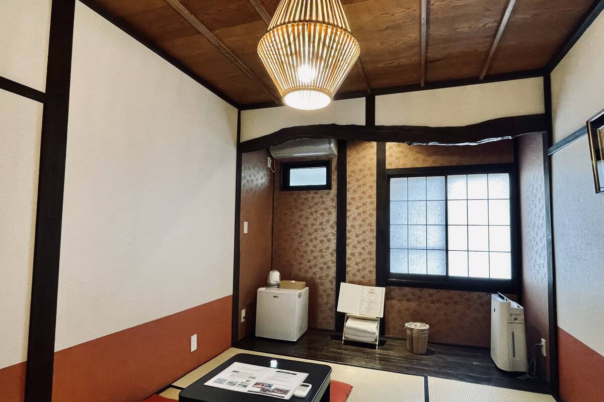 Ein traditioneller japanischer Raum mit Tatami-Böden, einem niedrigen Tisch mit einer Zeitung, einem Heizgerät, einem Mülleimer und einem Fenster mit Blick.
